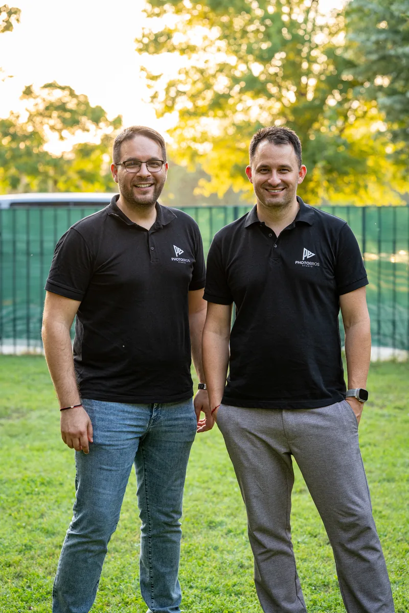 Two PhotoBros photographers in black studio polos on a lawn at sunset